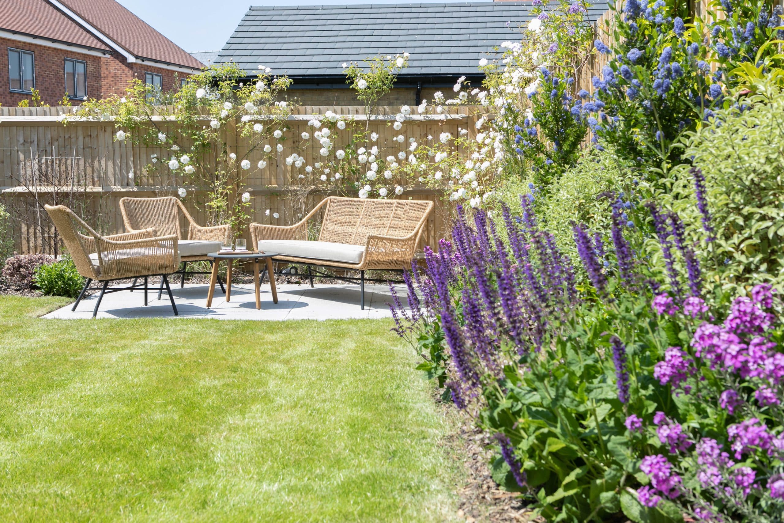 A garden with green grass and a patio with garden furniture on it. Colourful flowers border the garden.