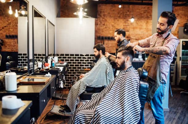A male barber cutting the hair of his client, wearing an apron.