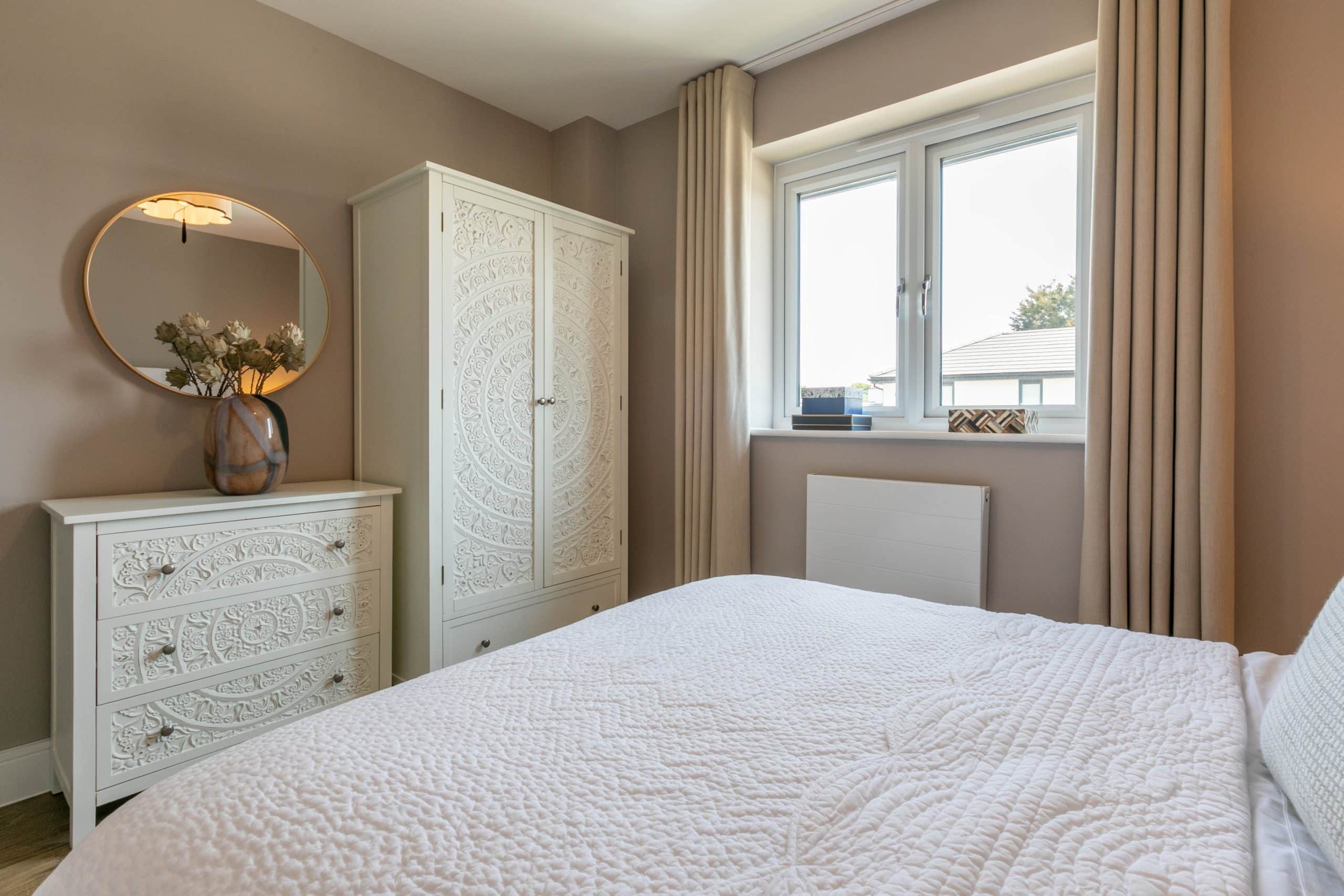 A bedroom with a bed facing a chest of draws and a wardrobe, with a window next to it.